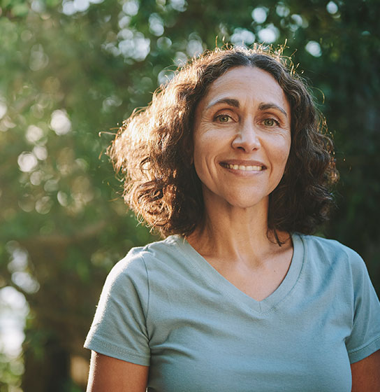 Smiling woman with curly brown hair in a light blue t-shirt outdoors, trees blurred behind her.