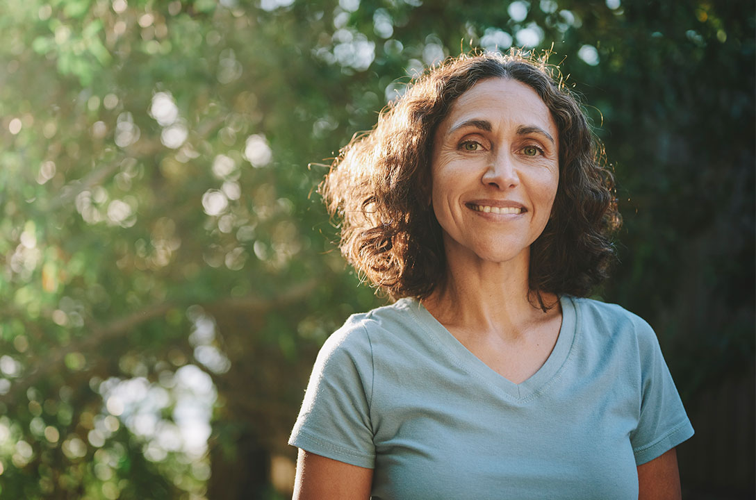 Smiling woman with curly brown hair in a light blue t-shirt outdoors, trees blurred behind her.