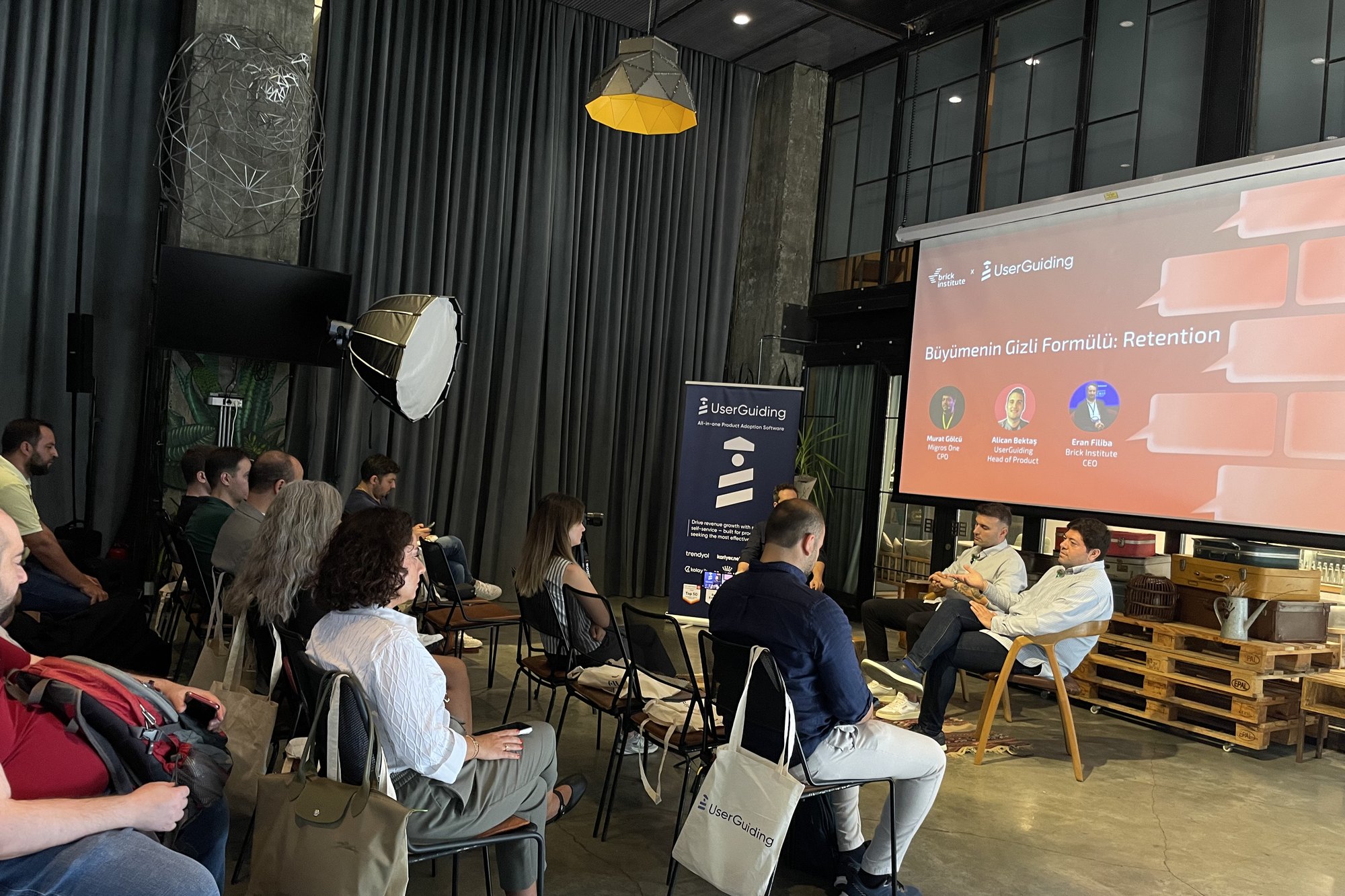 Fireside chat sahnesi: sağda iki panelist oturuyor, arkalarındaki ekranda Murat Gölcü, Alican Bektaş ve Eran Filiba'nın isimleri ve unvanları görünüyor, önde katılımcılar dinliyor