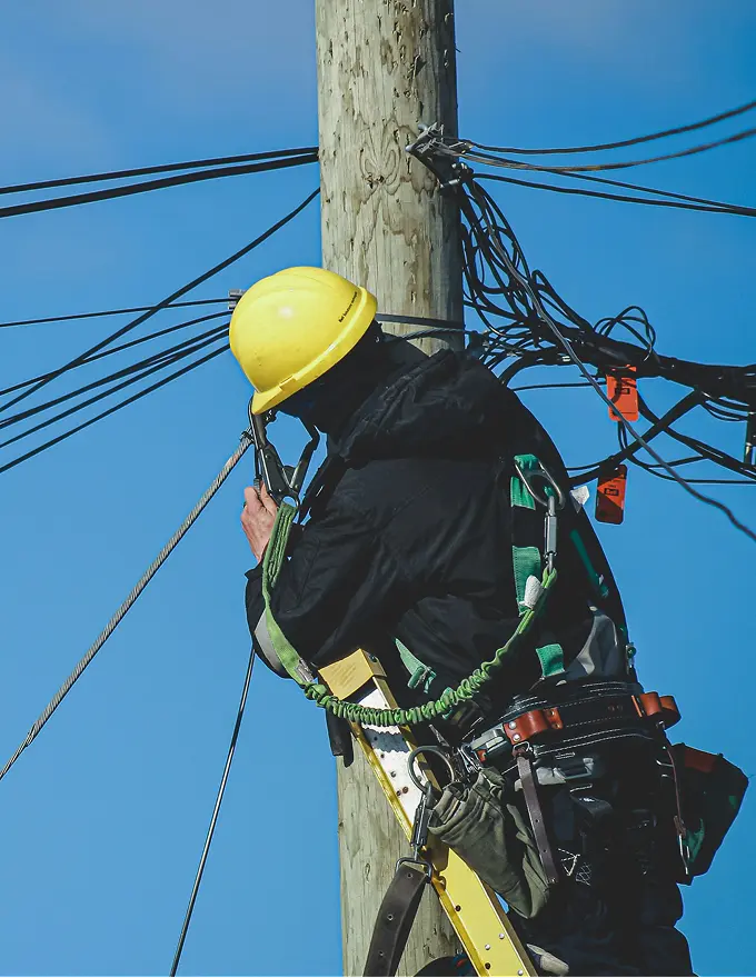 Telecom technician in a hard hat working on cables at the top of a utility pole.