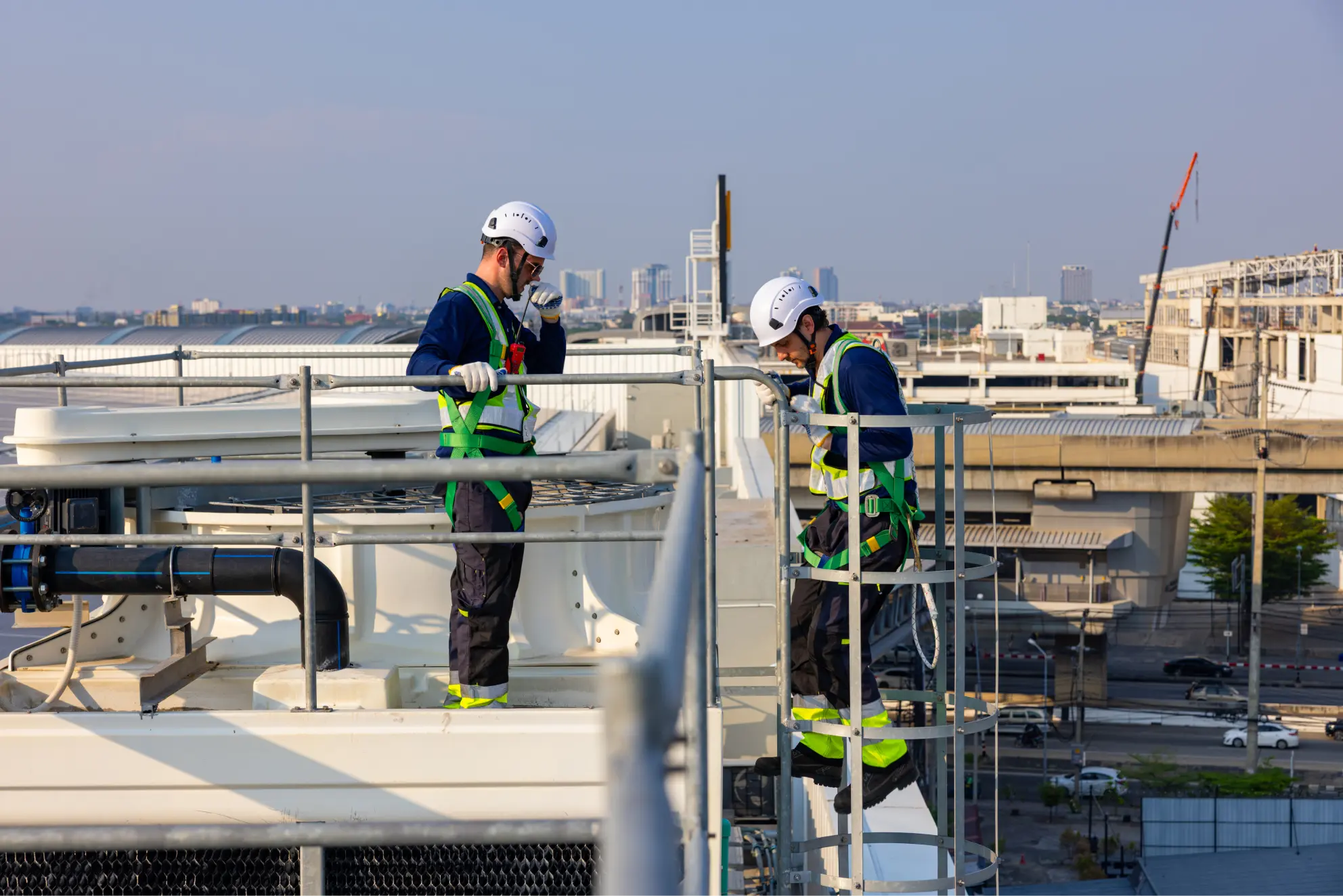 Two field technicians in safety harnesses and hard hats working on a rooftop cooling system.