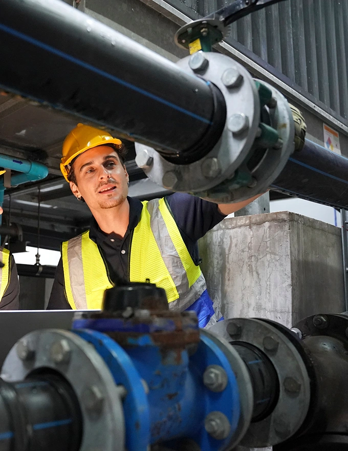 Water utility technician in a hard hat inspecting pipes and valves at a treatment facility.