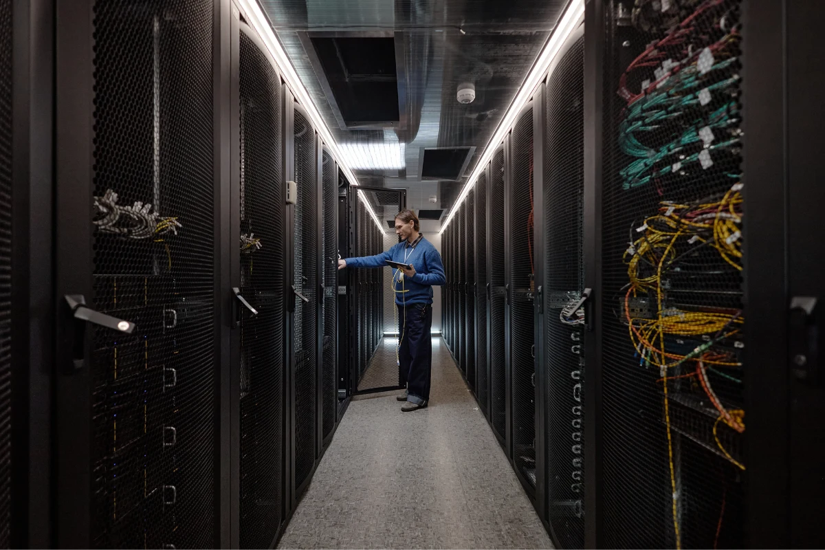 Technician with a tablet inspecting server racks and fibre cabling in a data center aisle.