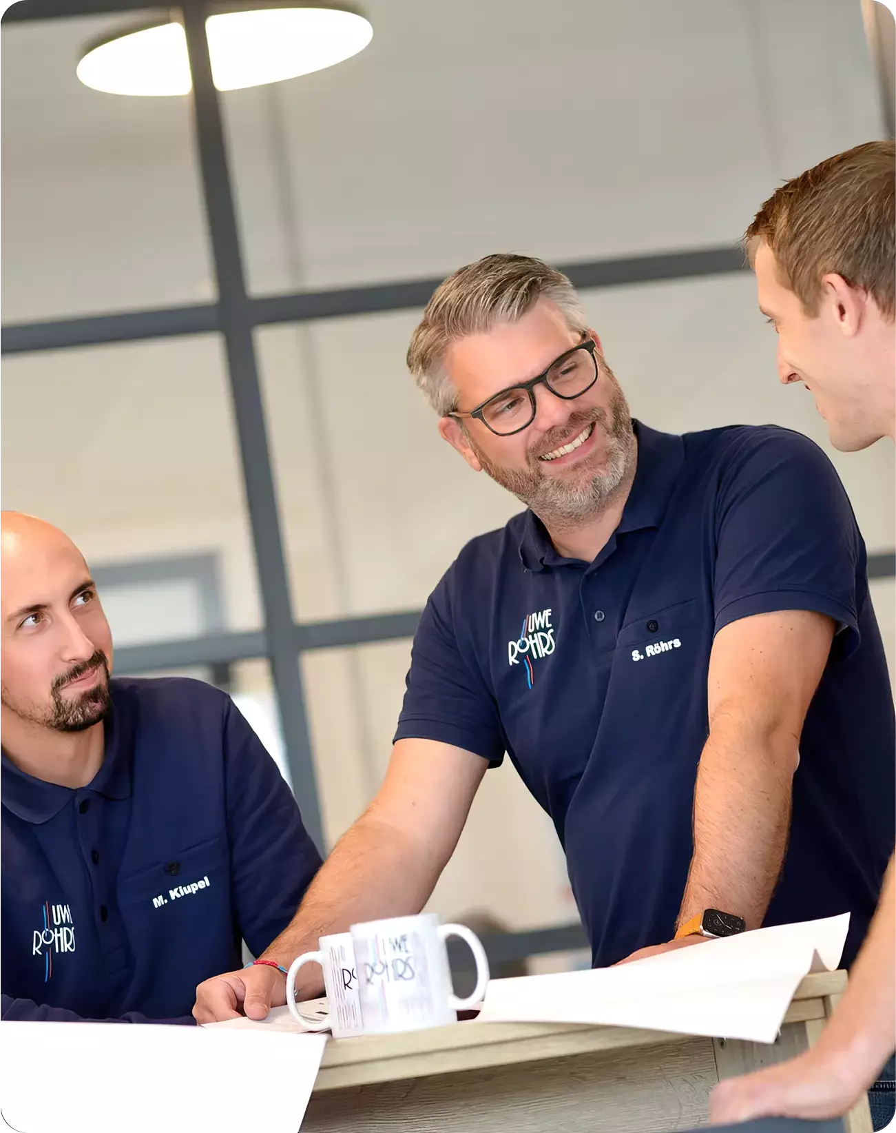 Drei Männer in blauen Poloshirts diskutieren bei einem Tisch mit Kaffeetassen und Papierplänen.
