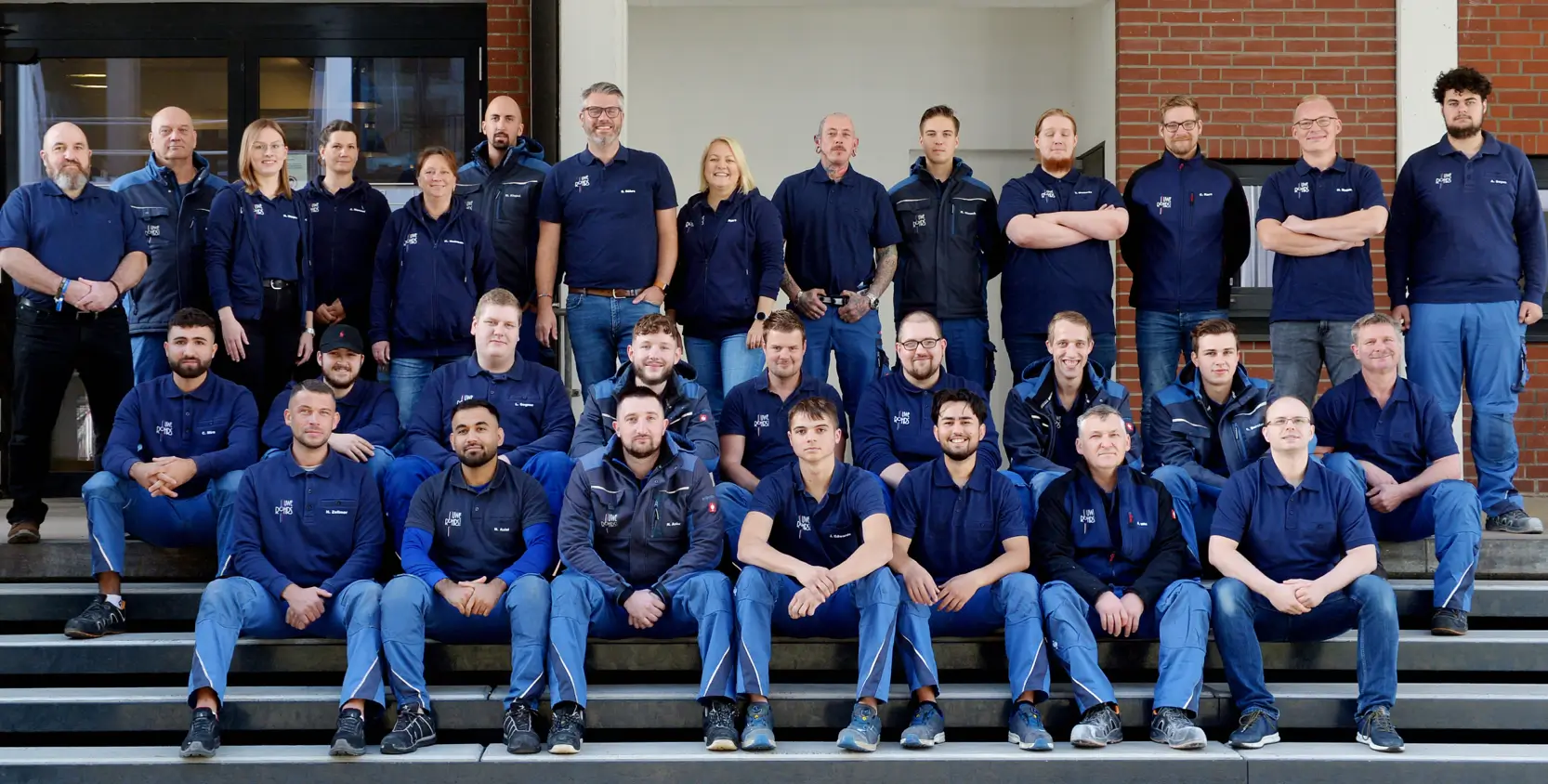 Gruppenfoto von 30 Mitarbeitern in blauen Arbeitskleidungen, vor einem Gebäude mit roten Ziegeln.