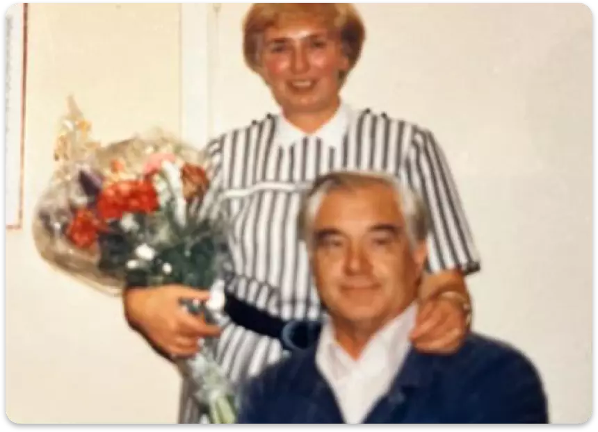 Ein älteres Paar, Frau mit Blumenstrauß und Mann in blauer Jacke, sitzt zusammen in Innenraum.