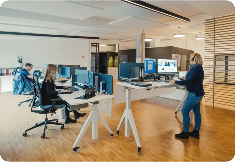 Büro mit zwei sitzenden Personen an Computern und einer stehenden Frau mit Headset am Stehpult.