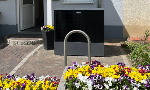 Wärmepumpe vor einem Haus mit bunten Blumen und einem offenen weißen Türrahmen.