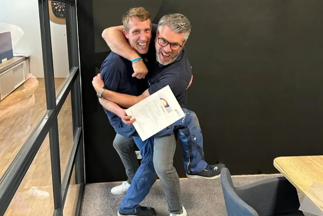 Zwei Männer umarmen sich lachend, einer hält ein Dokument in einem modernen Büro.