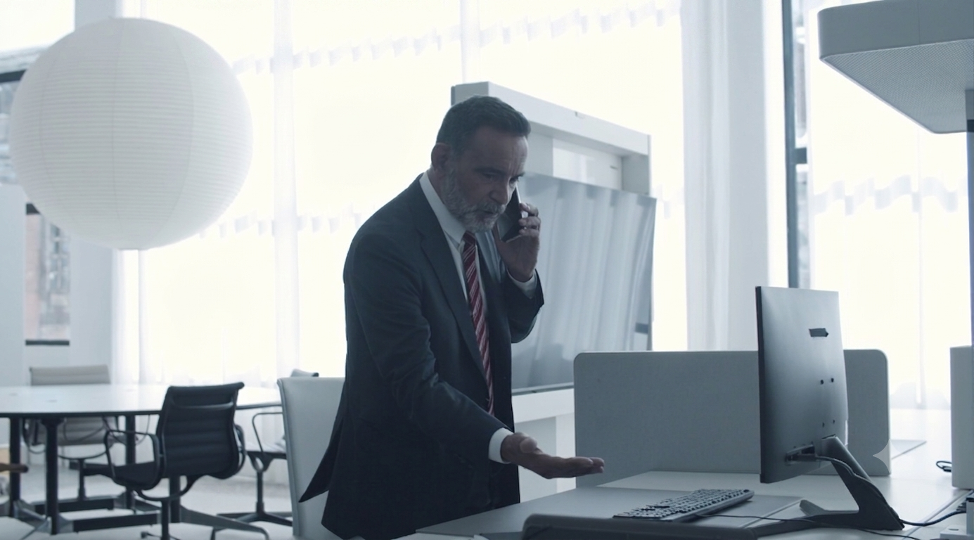 Man in suit talking on phone looking at his palm over a desk with computer and keyboard in office.