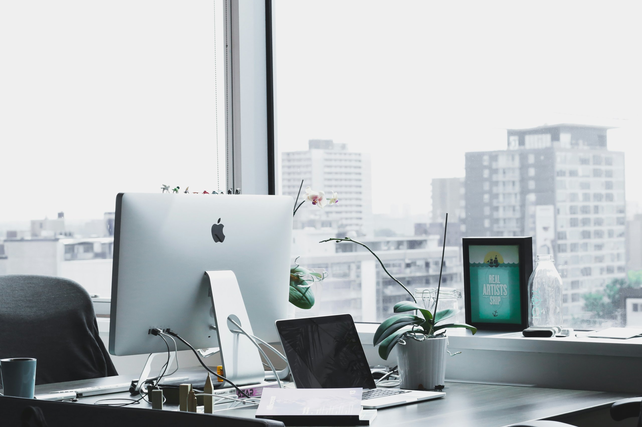 Office desk with Apple iMac, laptop, potted orchid, and city view through window.