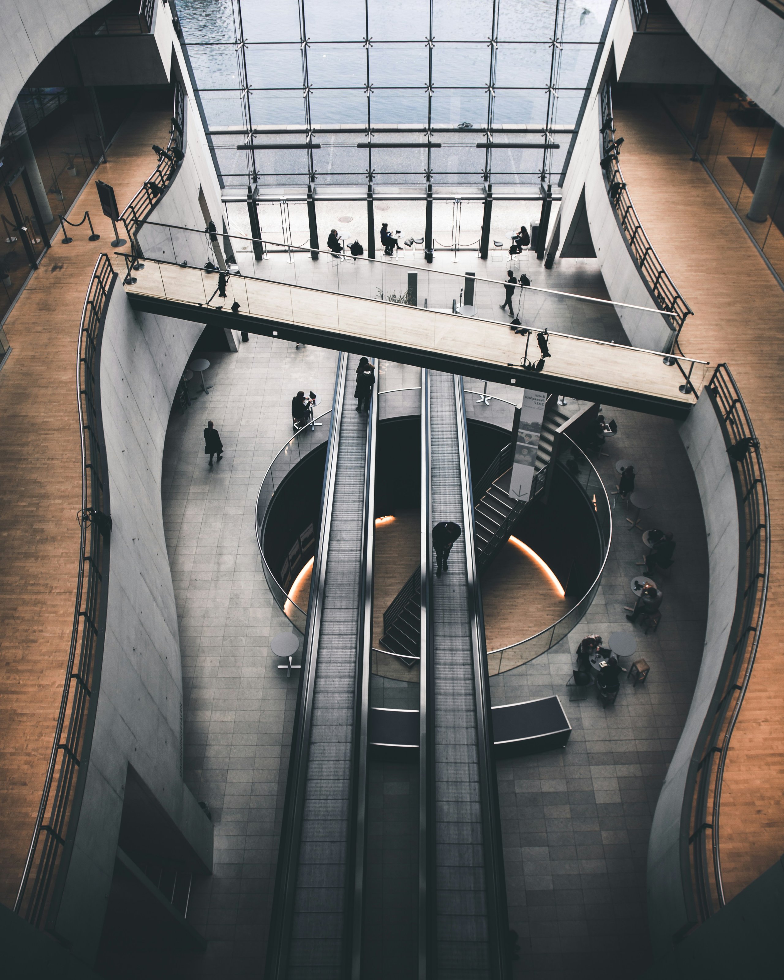 Modern building interior with escalators, circular stairway, wooden bridge, and people walking and sitting.