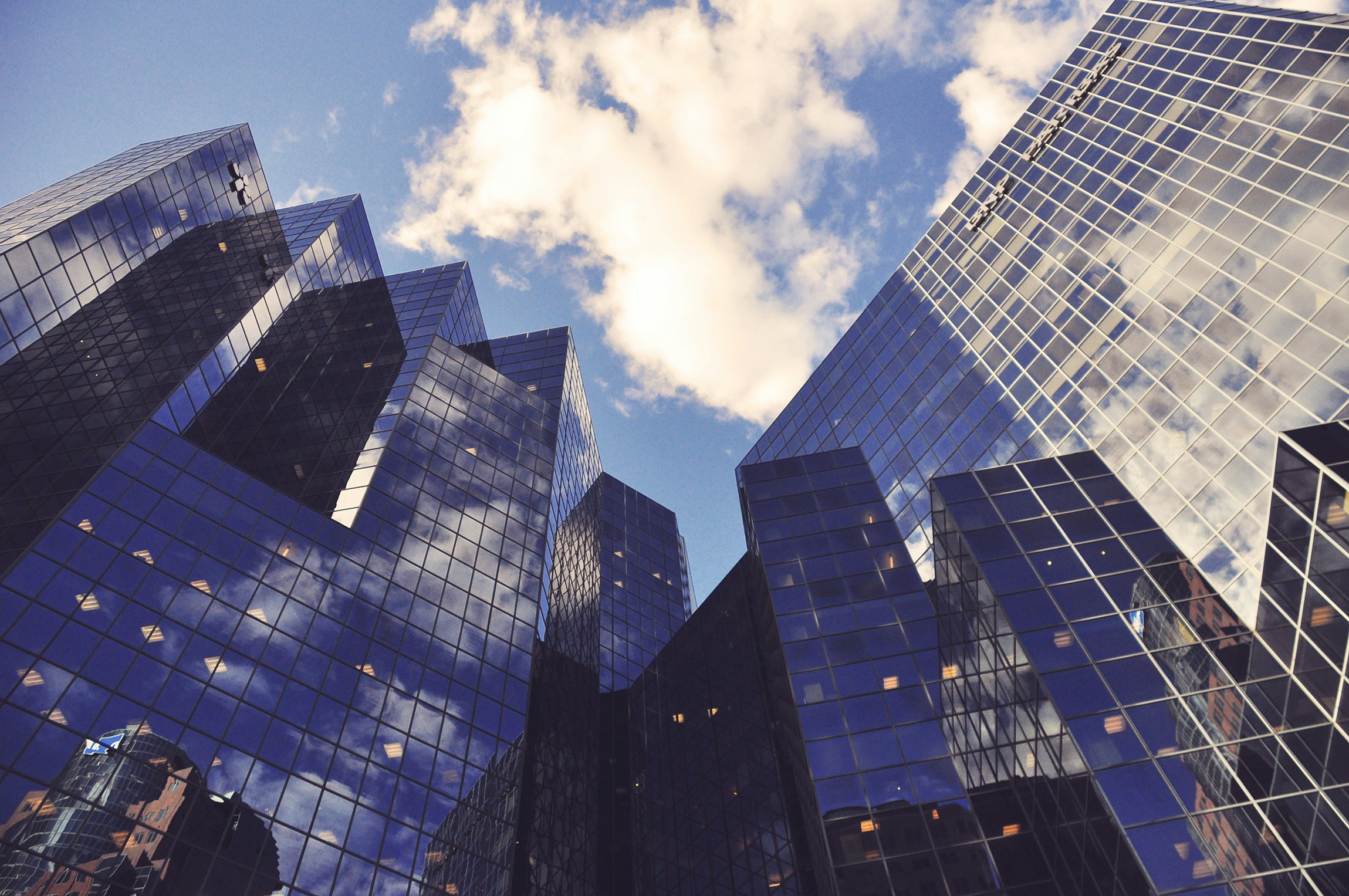Tall glass skyscrapers reflecting clouds and blue sky from a low angle perspective.