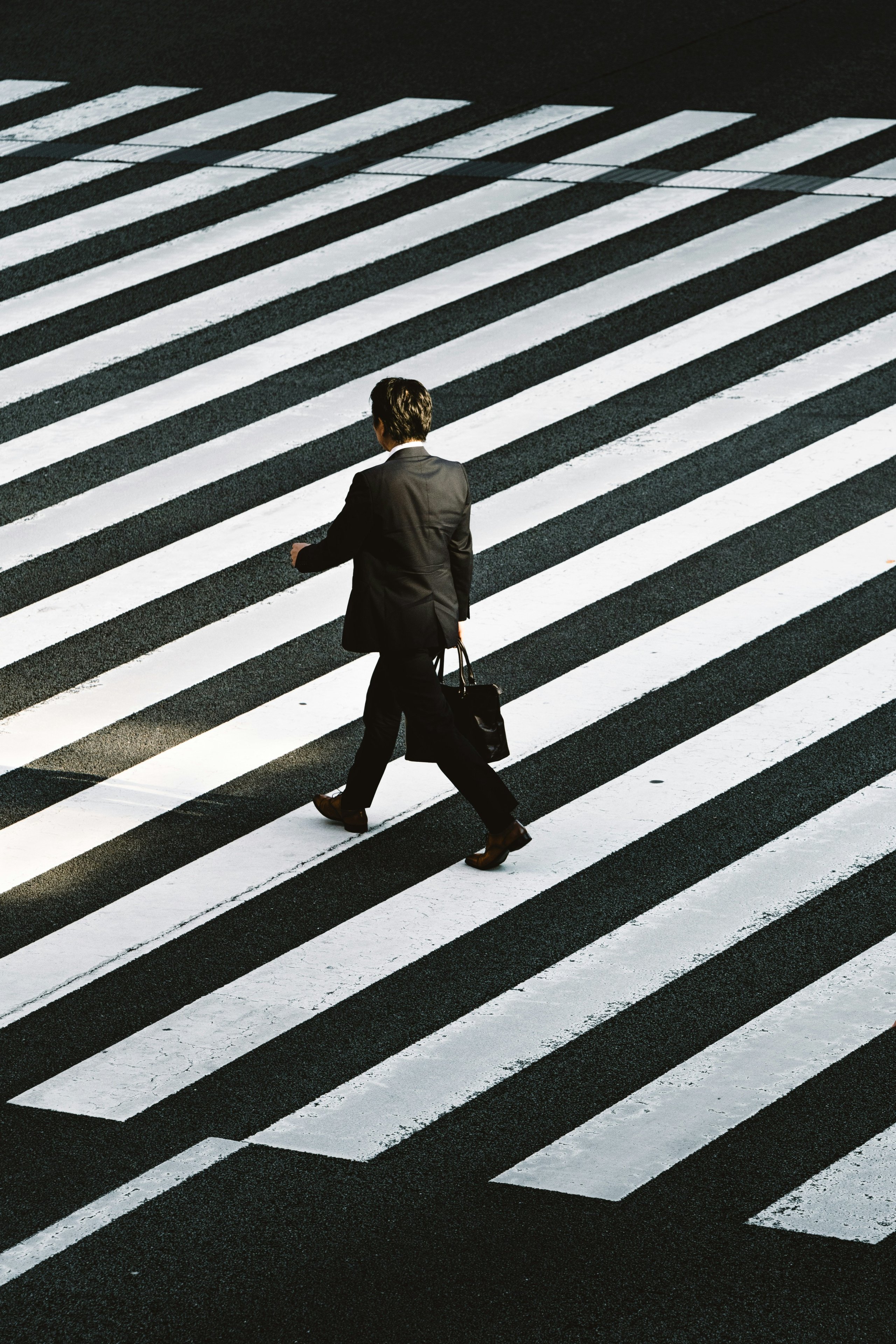 Person in a black suit crossing a striped pedestrian crosswalk while carrying a black bag.