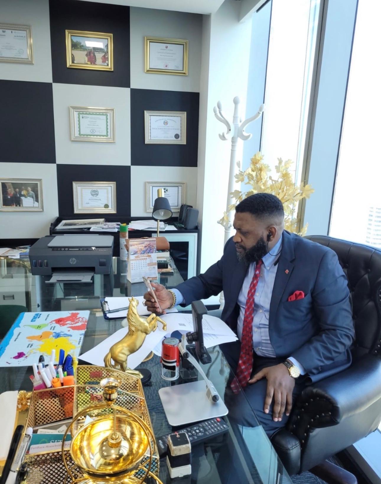 Man in suit sitting at a glass office desk using a smartphone with certificates on the wall behind him.