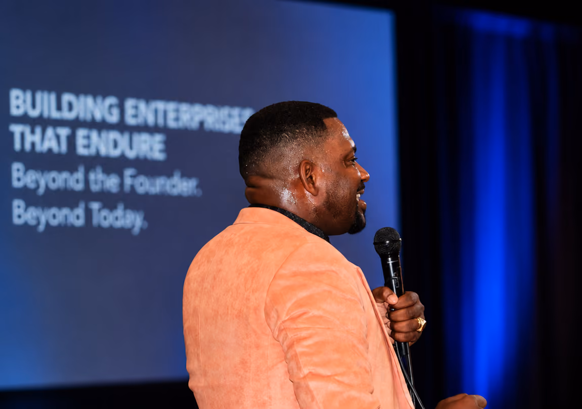 Man in peach jacket speaking into microphone with presentation slide in background.