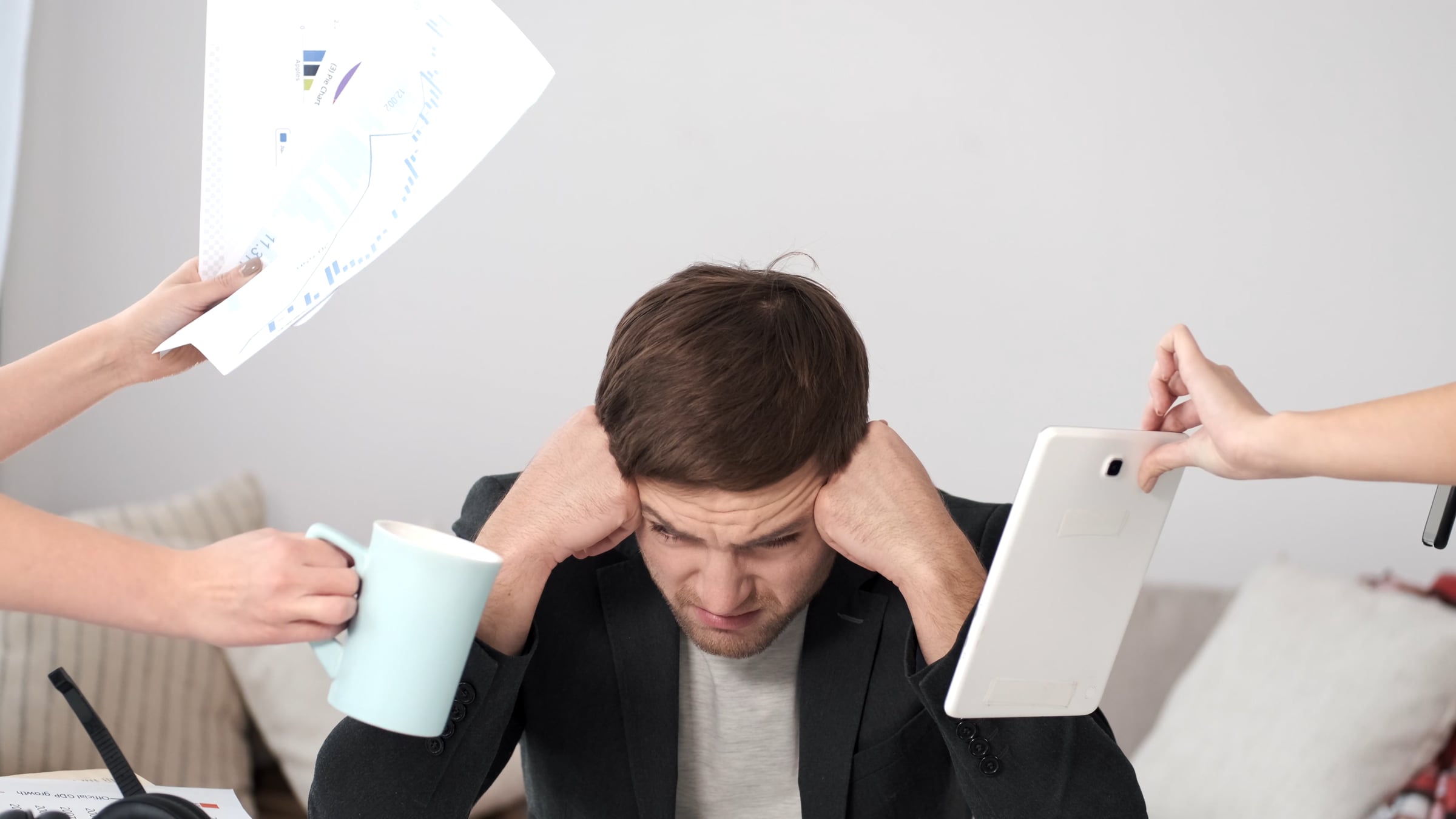 Stressed man holding his head as hands offer papers, a mug, and a tablet around him.