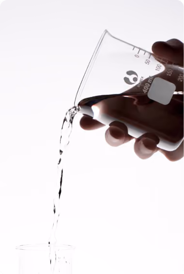 Hand pouring clear liquid from a glass beaker into another container against a white background.