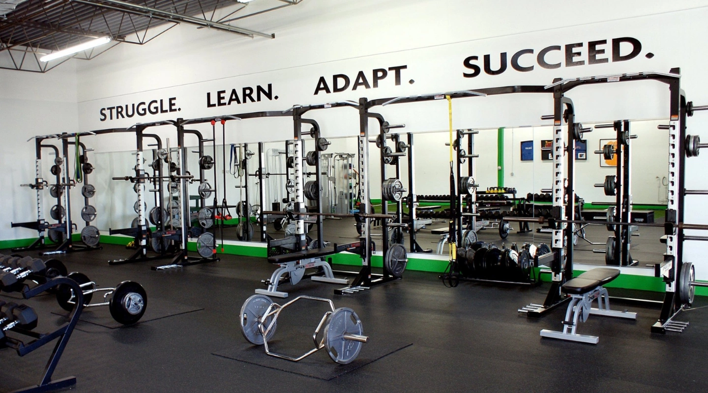 An indoor gym featuring multiple squat racks, weight benches, barbells, and dumbbells on a rubber floor, with the motivational phrase "STRUGGLE. LEARN. ADAPT. SUCCEED." painted in large letters across the back wall.