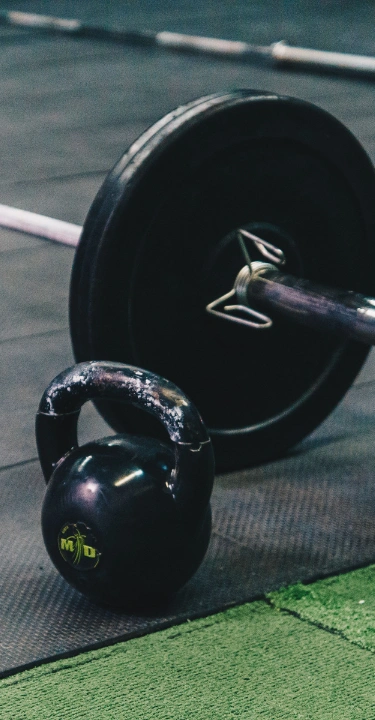 A black kettlebell sits on a dark rubber gym floor next to a loaded barbell with weight plates.