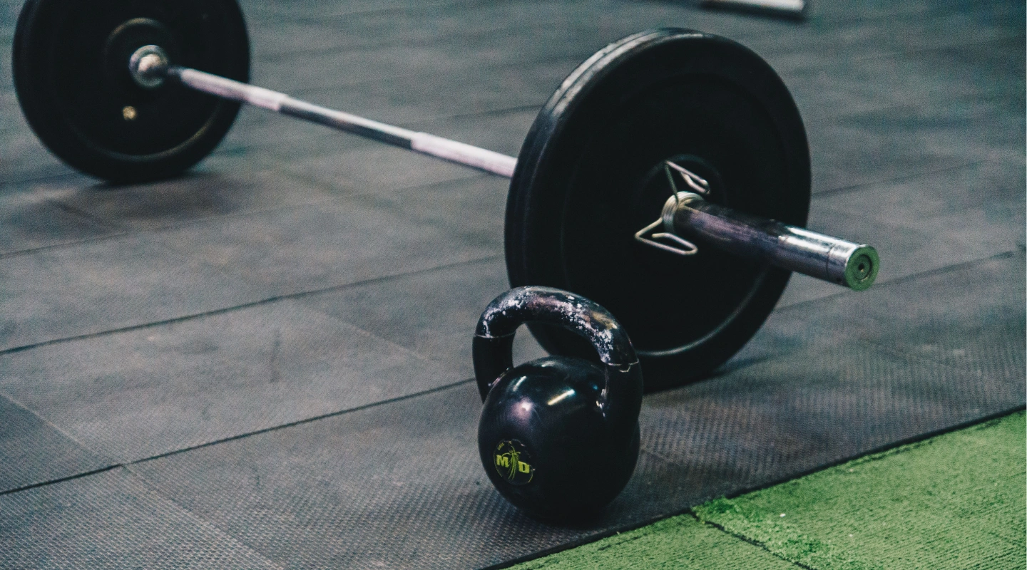 A black kettlebell sits on a dark rubber gym floor next to a loaded barbell with weight plates.