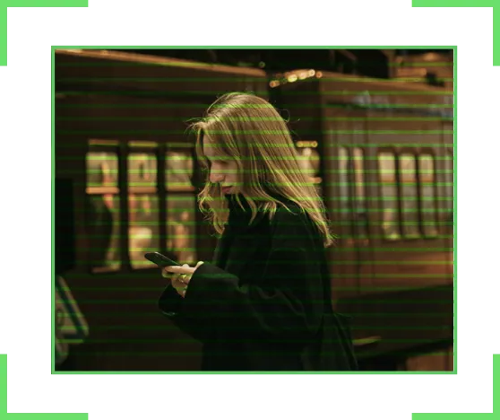 Woman in dark coat looking at her phone on a subway platform with train cars behind her.