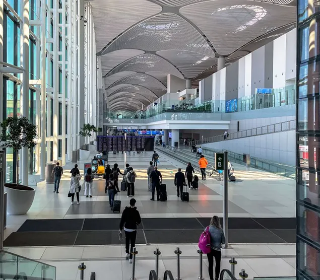 People walking with luggage in a spacious, modern airport terminal with high ceilings and large windows.