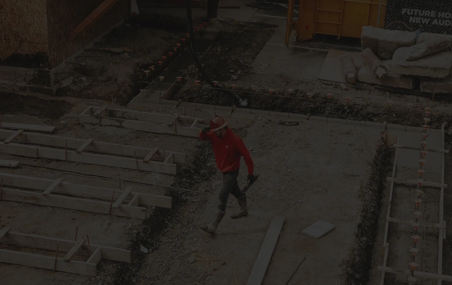 Construction worker in red hoodie and helmet walking on a concrete foundation site.