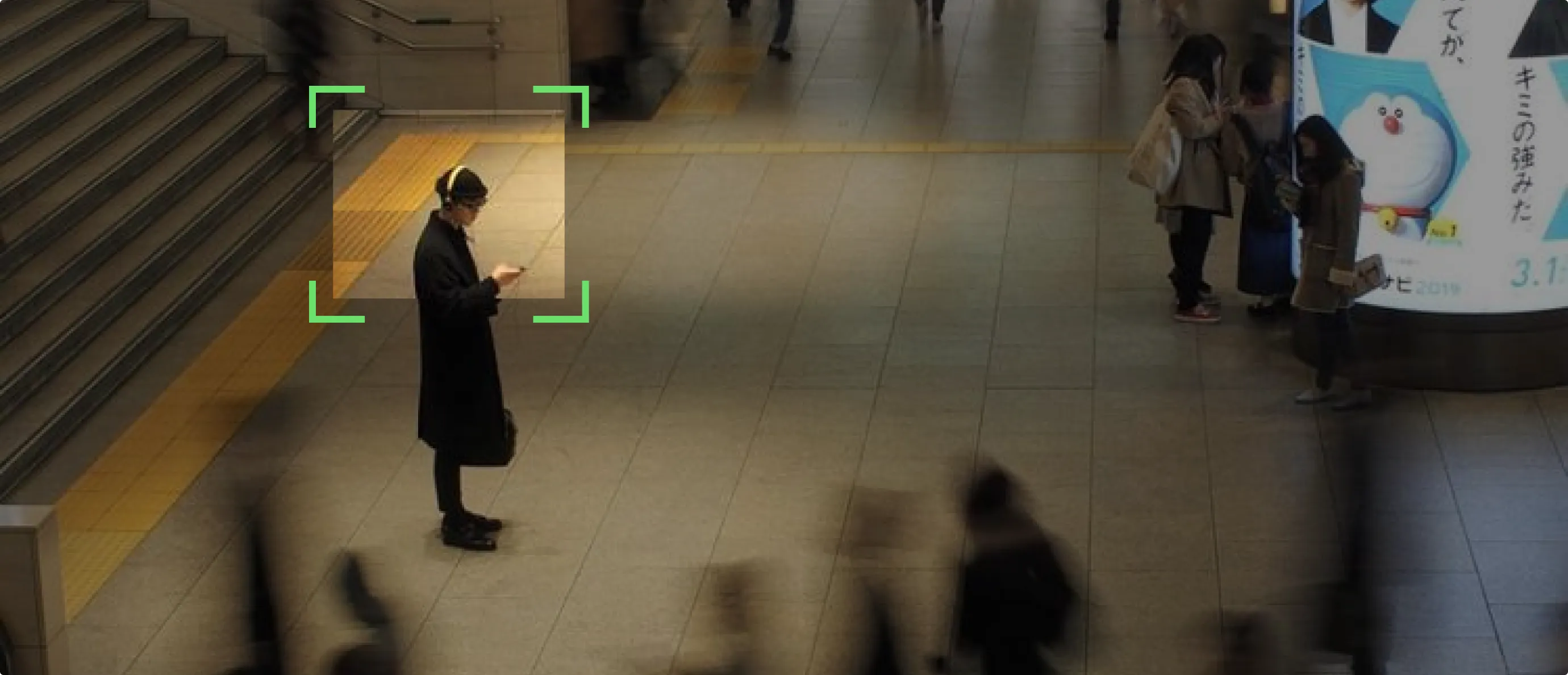 Person in black coat and headphones looking at phone in a busy indoor public space with blurred people.