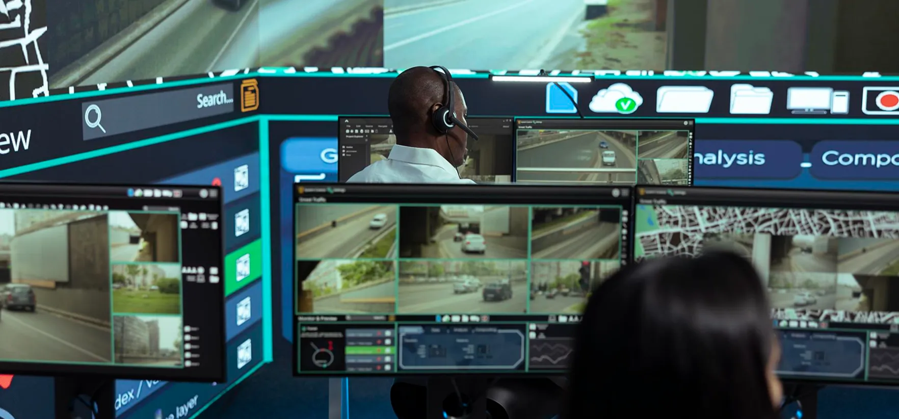 Two people monitoring multiple traffic camera feeds on computer screens in a control room.