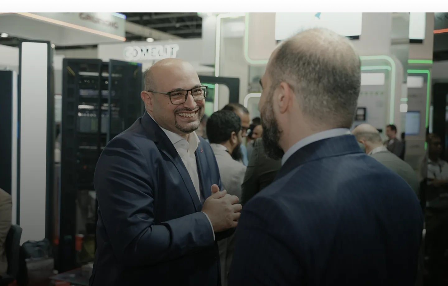 Two men in suits talking and smiling at a busy tech event or conference.