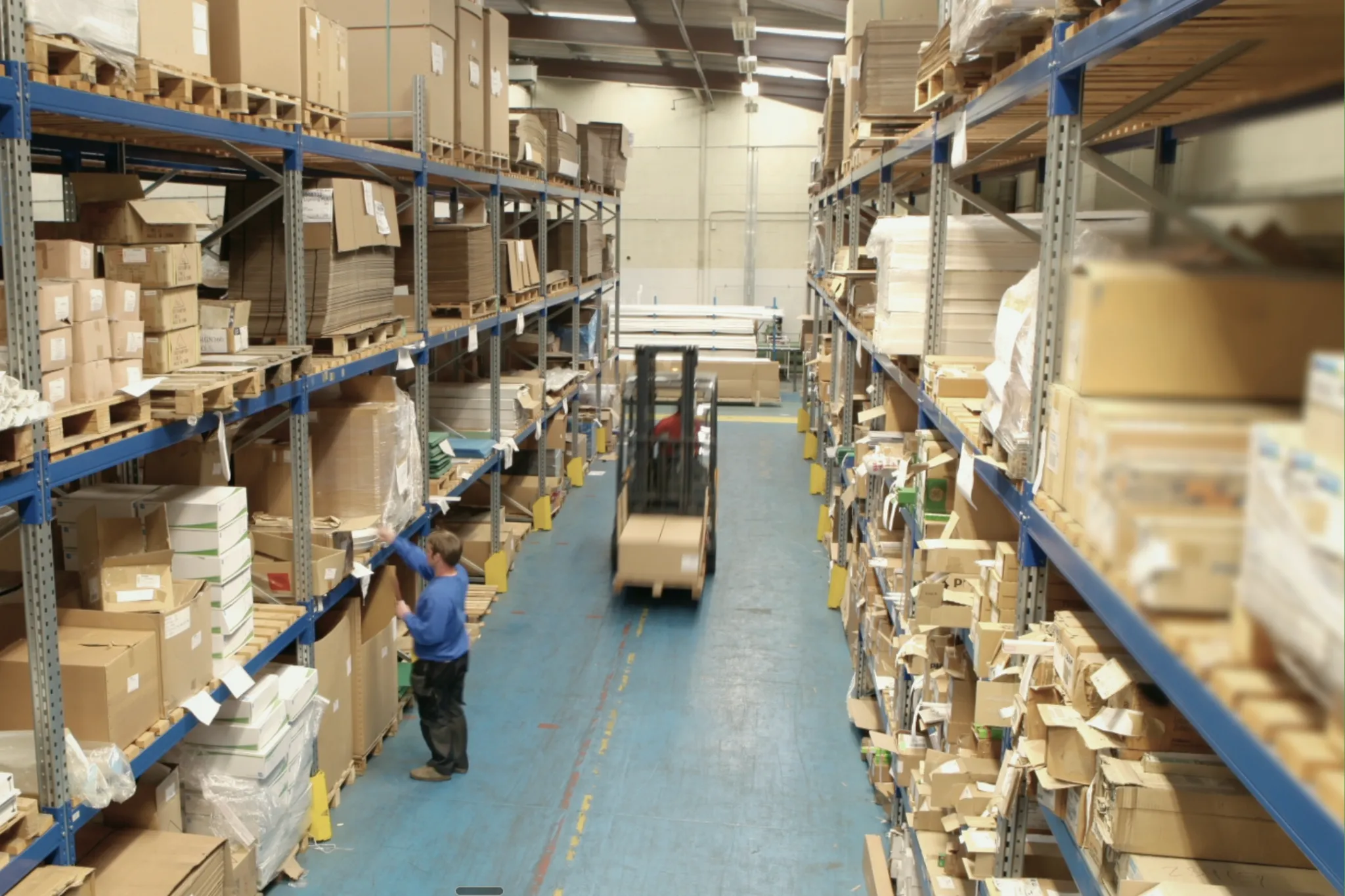 Warehouse aisle with shelves full of boxes, a worker organizing items, and a forklift carrying a pallet.