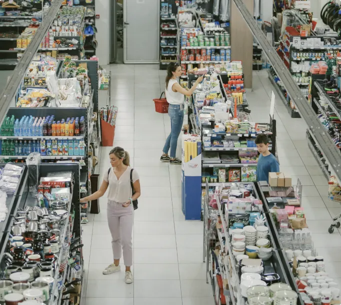 Customers browsing various kitchenware and household items in supermarket aisles with well-lit white tile floors.