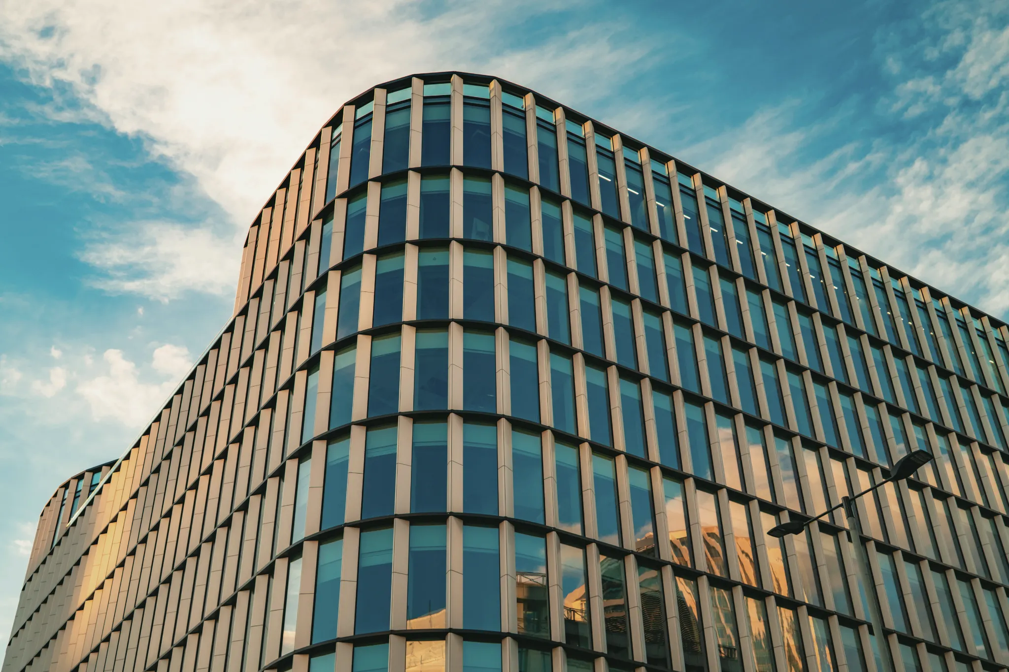Modern office building with curved glass windows reflecting sunset and blue sky with clouds.