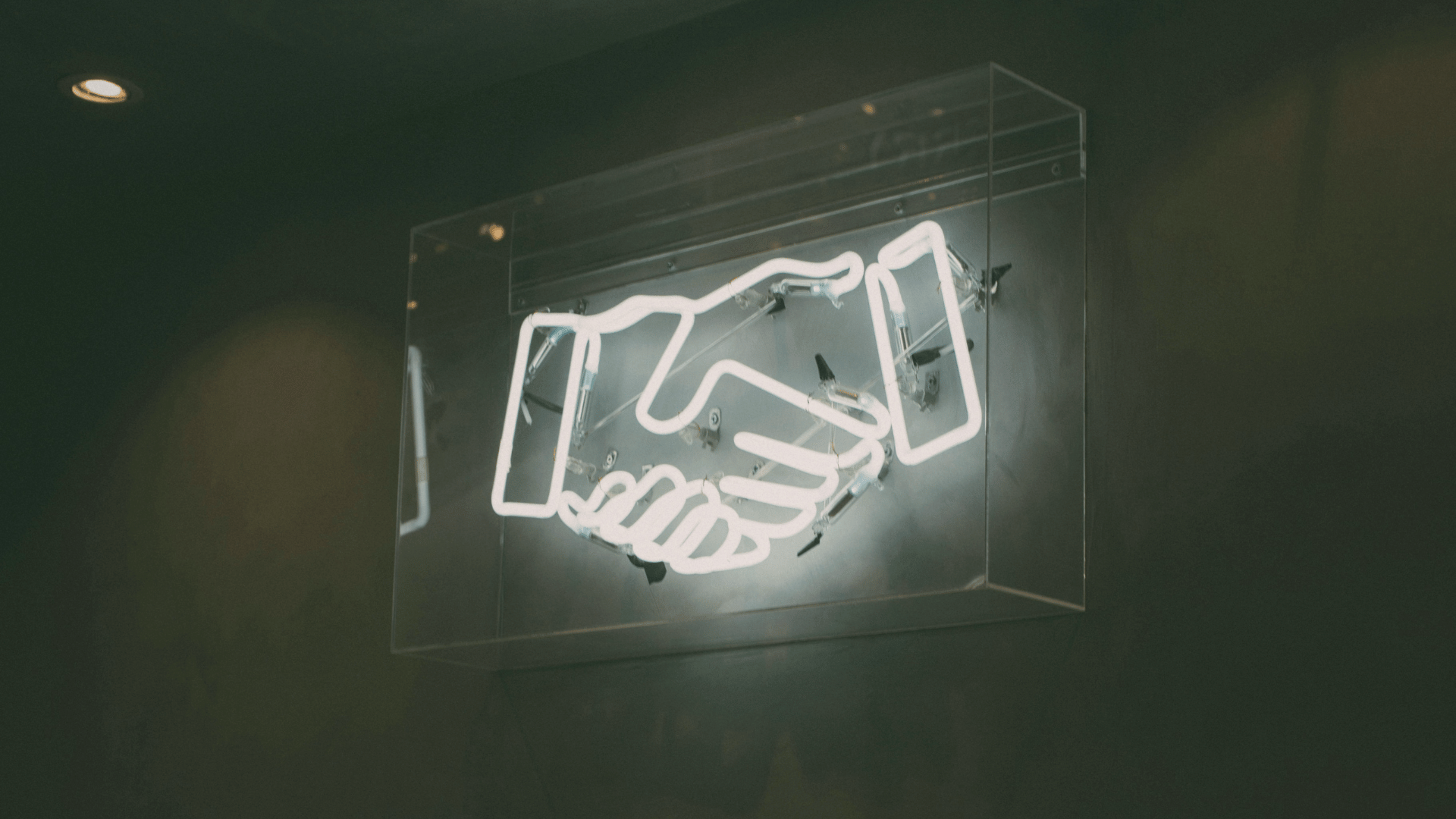 White neon light sign shaped like two hands shaking inside a clear box on a dark wall.