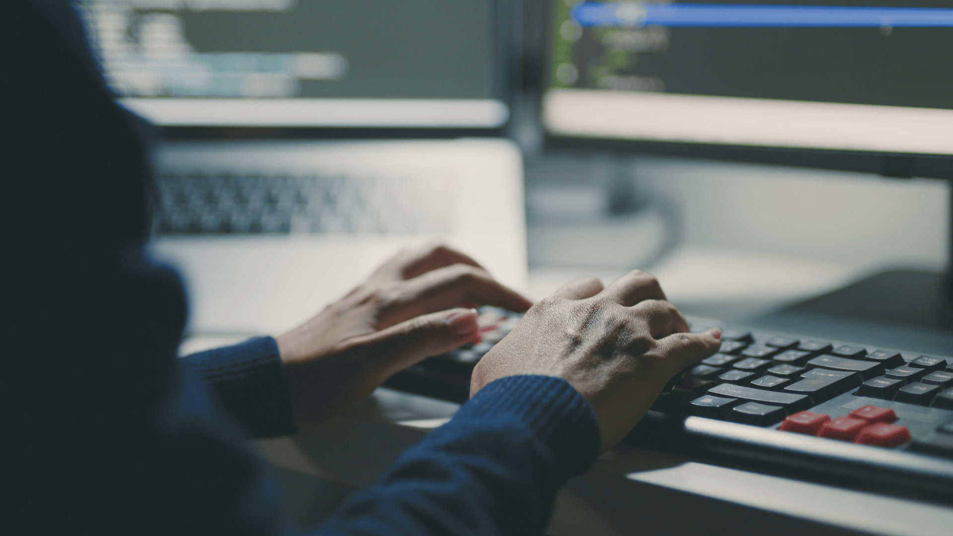 Person typing on keyboard with code displayed on computer screens in the background.