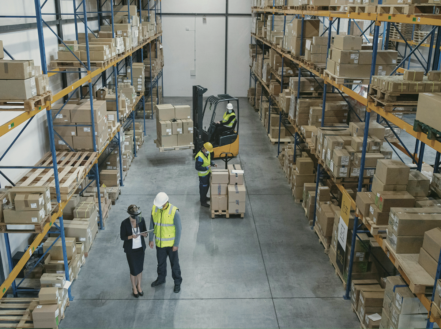Warehouse workers wearing safety gear with forklift moving boxes on pallets amid tall shelving.