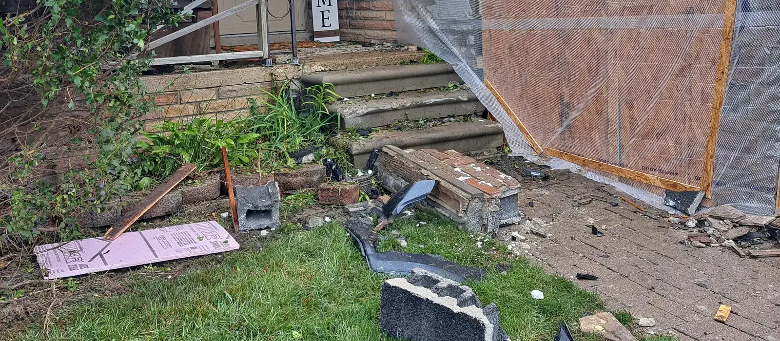 Broken concrete block and debris on the lawn beside the boarded wall where a vehicle hit the house
