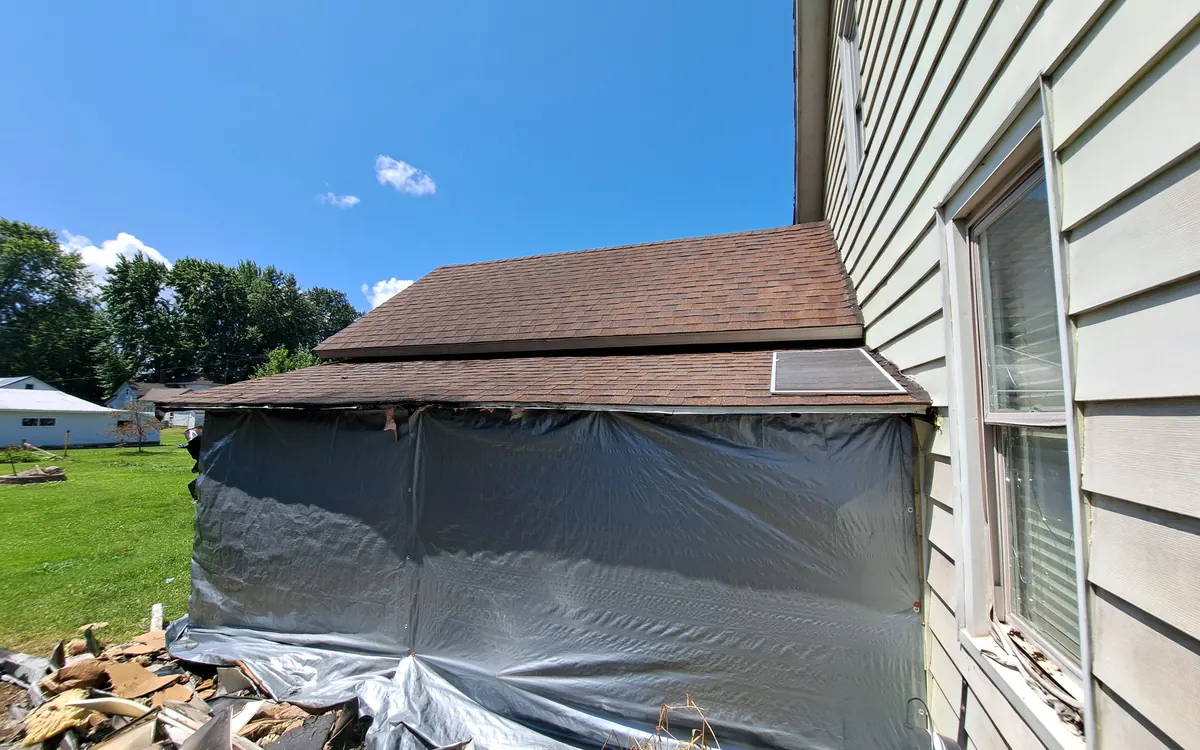 Side of a house wrapped in plastic sheeting after a vehicle fire, with burned debris on the ground