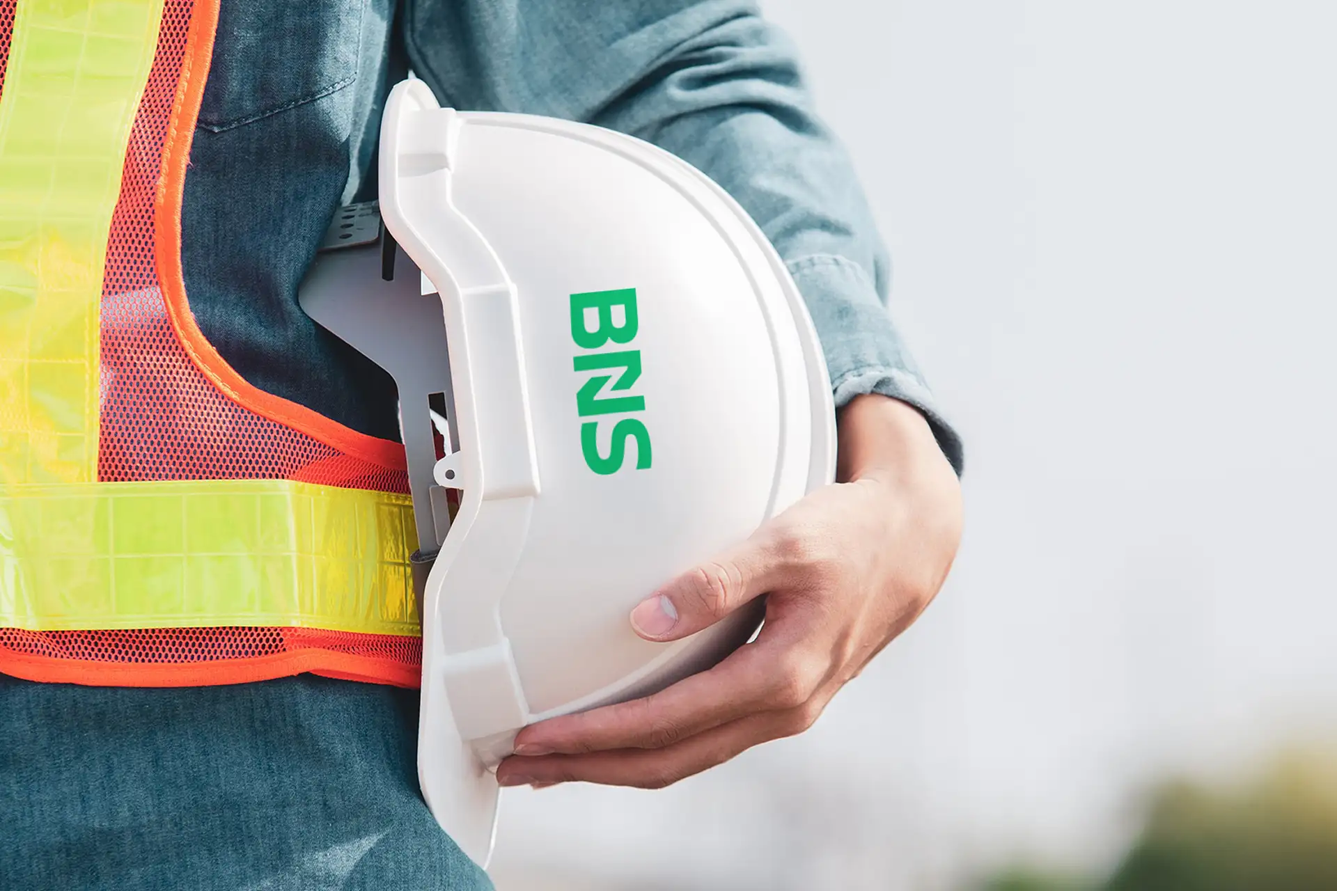 Persoon houdt een witte bouwhelm met groene letters BNS en draagt een fluoriserend veiligheidsvest.