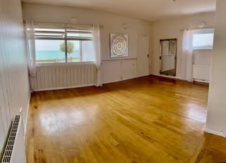 Empty room with wooden floor, large window, and mirror reflecting the sea view outside.