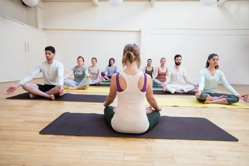 A group of people sitting on yoga mats, meditating in a light-filled studio.