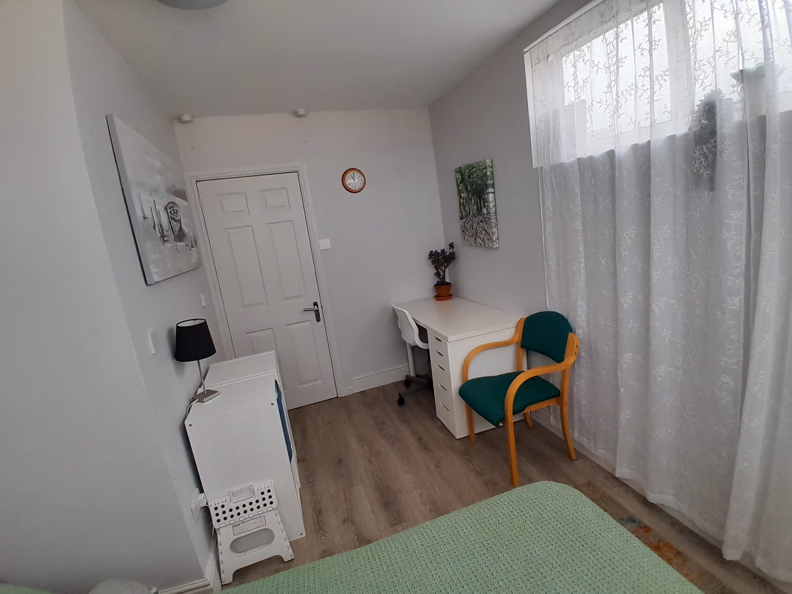A small room with a desk, chair, and white curtains beside a bed.