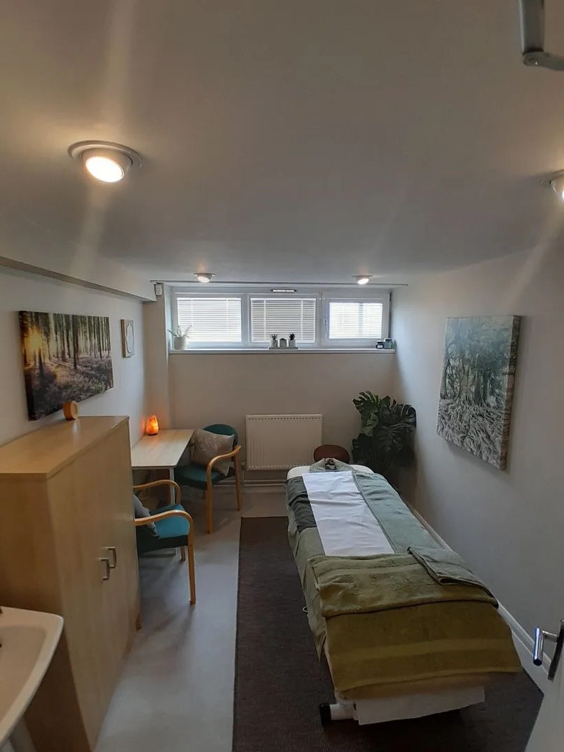 A massage table with a folded towel is in a small, well-lit room with paintings and plants.