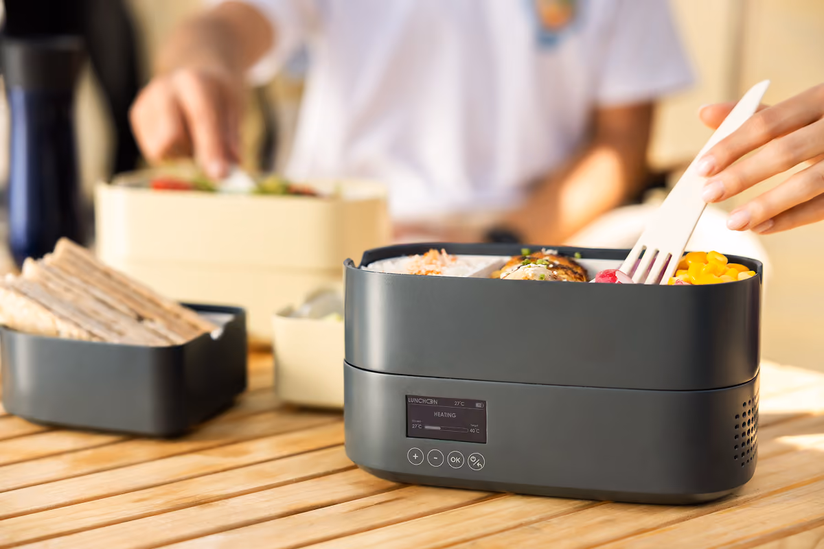 Black electric lunch box with grilled chicken, ramen noodles, sliced boiled eggs, seaweed, and side dish with rice.