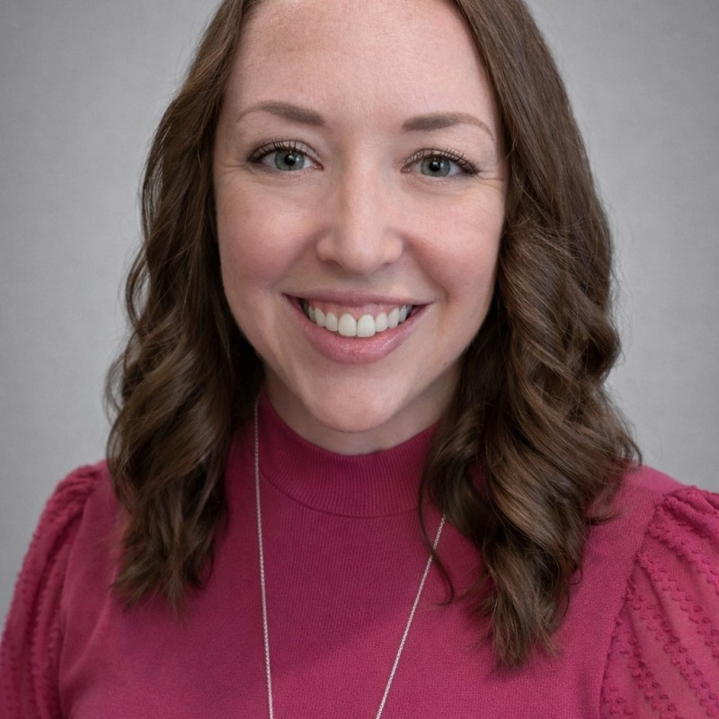 Smiling woman with wavy brown hair wearing a rose-colored top against a gray background.