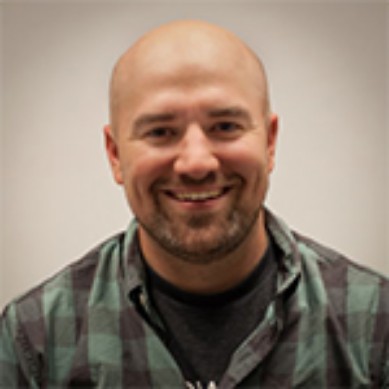 Smiling bald man with beard wearing a green and black plaid shirt against a neutral background.