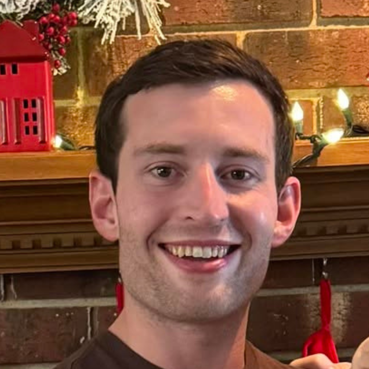 Smiling young man with short dark hair in front of a brick wall decorated with holiday lights and a red lantern.