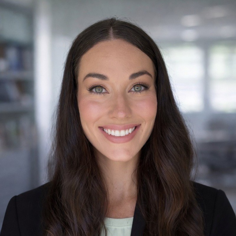 Smiling woman with long dark hair and green eyes wearing a black blazer and light top indoors.