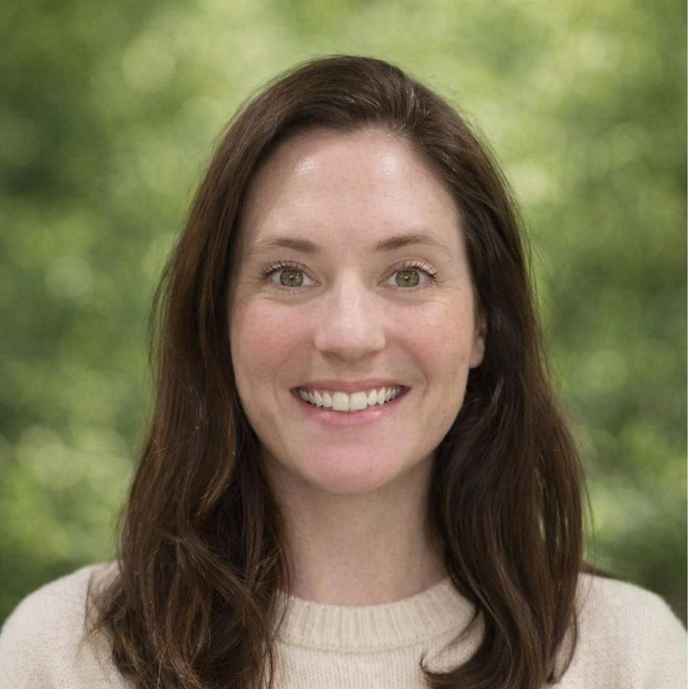 Lizzie has long brown hair wearing a cream sweater against a blurred green background.